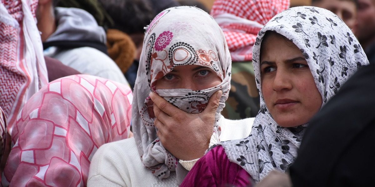 A crowd of people with two women facing the camera. These women are wearing traditional clothing for Syrian women.